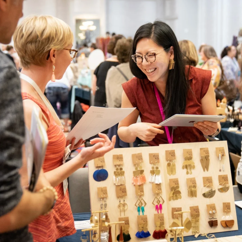 Woman showing earrings to another woman at a trade show or similar event.