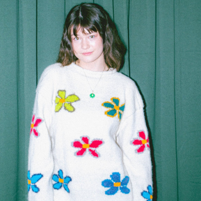 A young woman with dark hair wearing a white fuzzy knit sweater decorated with colorful crocheted flowers, standing in front of a dark green curtain.
