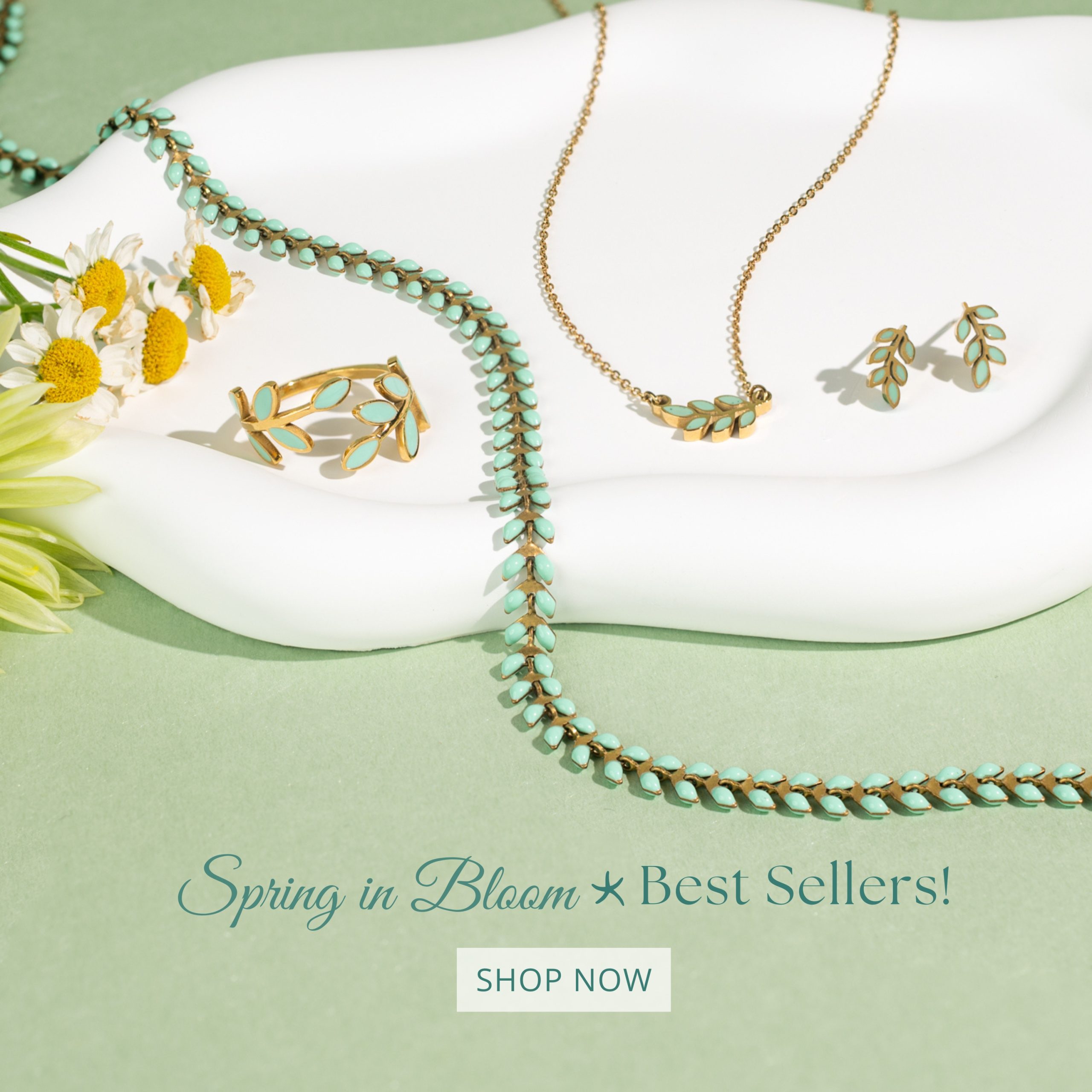 Gold jewelry with light blue leaf accents, including a ring, necklace, and earrings, is displayed on a white tray with daisies on a green background. Text reads Spring in Bloom ★ Best Sellers! SHOP NOW.