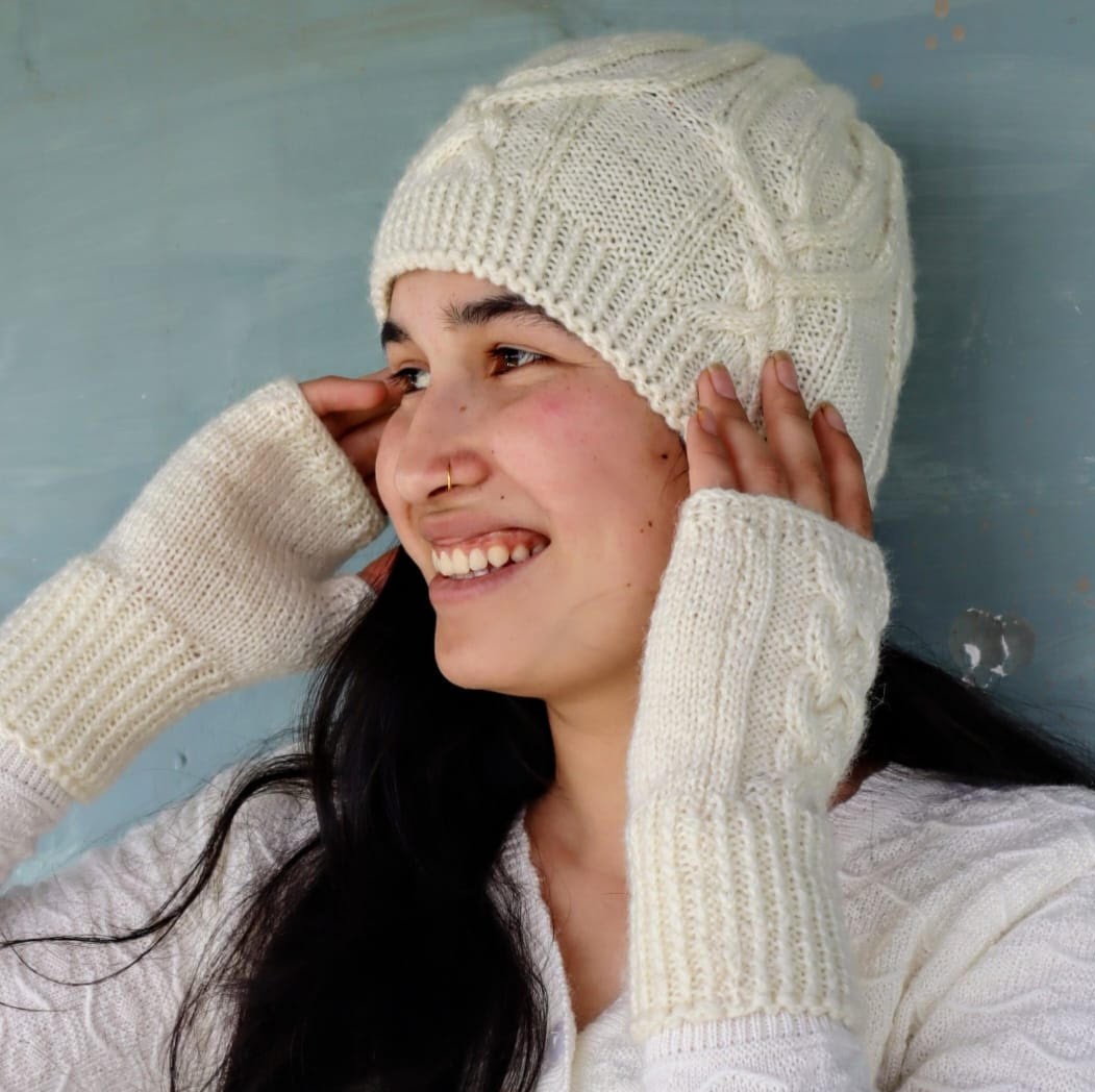 A smiling woman wearing a cream-colored knitted hat, matching fingerless gloves, and a white sweater stands against a blue background, touching the sides of her hat with both hands.