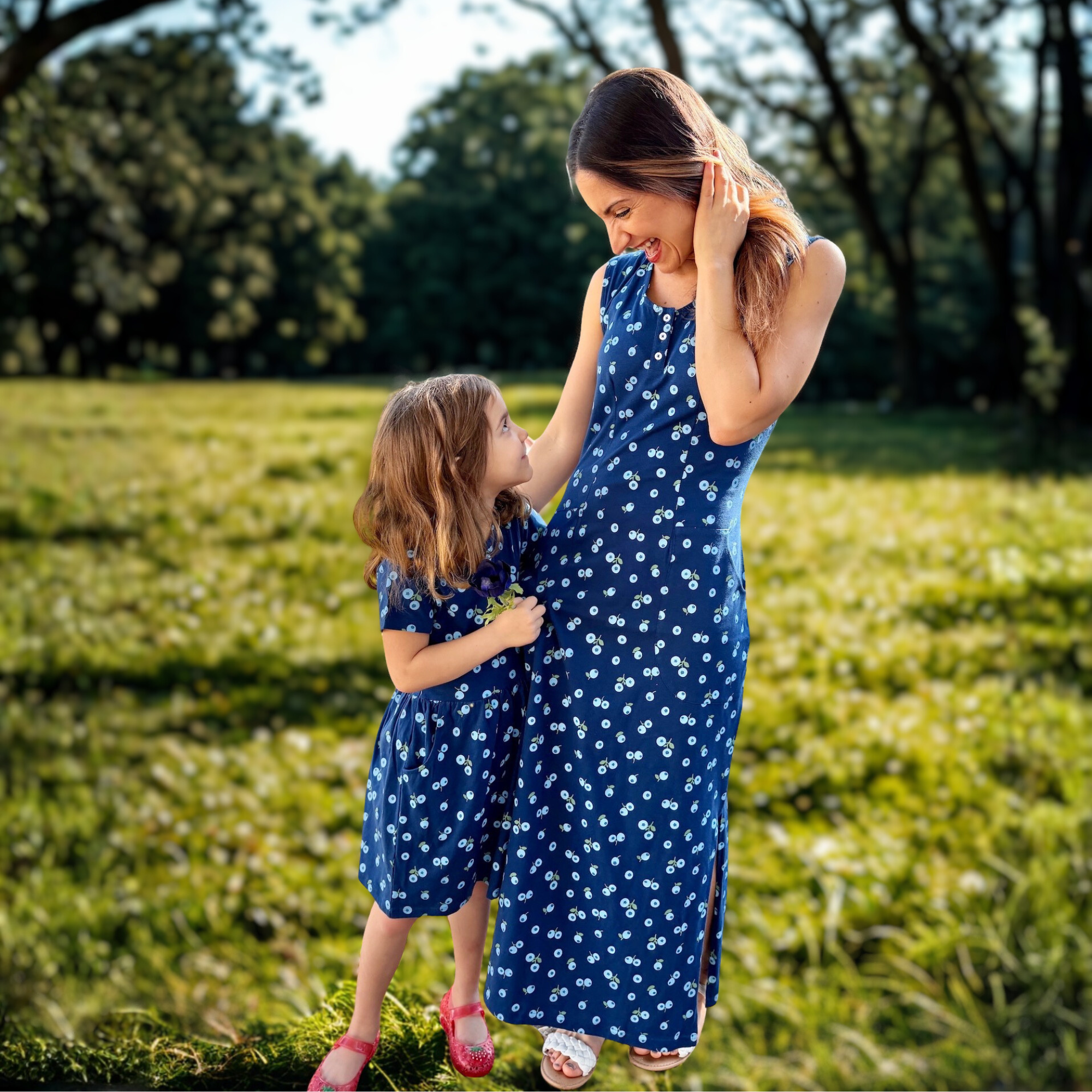 A woman and a young girl, both wearing matching blue dresses with white patterns, smile and look at each other while standing in a sunlit grassy field with trees in the background.