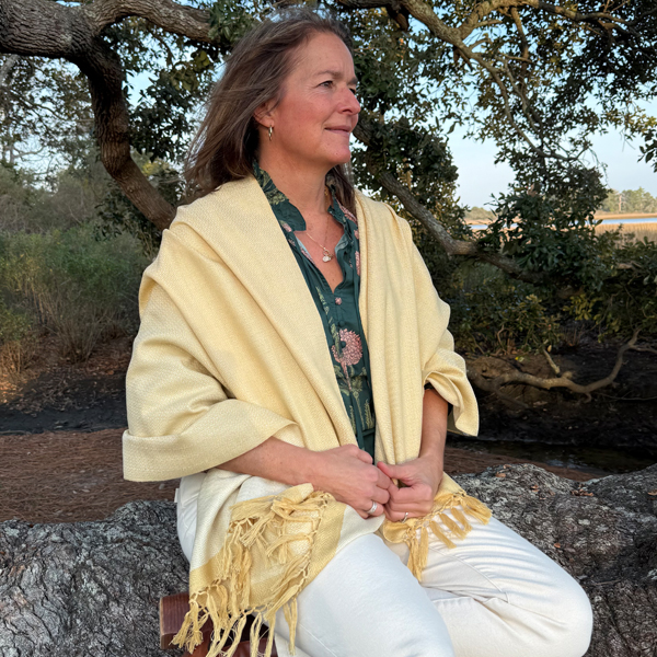 A woman with long hair, wearing a green floral shirt, white pants, and a light yellow shawl, sits on a large tree branch outdoors, smiling and looking to the side. Trees and greenery are in the background.