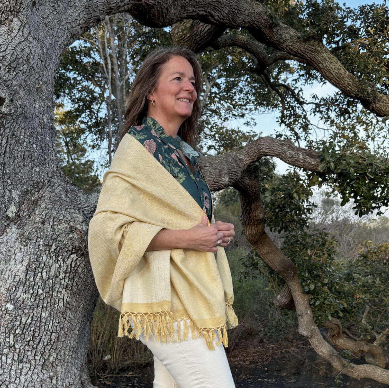 A woman stands outdoors by a large, twisted tree, smiling. She wears a yellow shawl over a green patterned shirt and white pants. The background is filled with greenery and leaves.