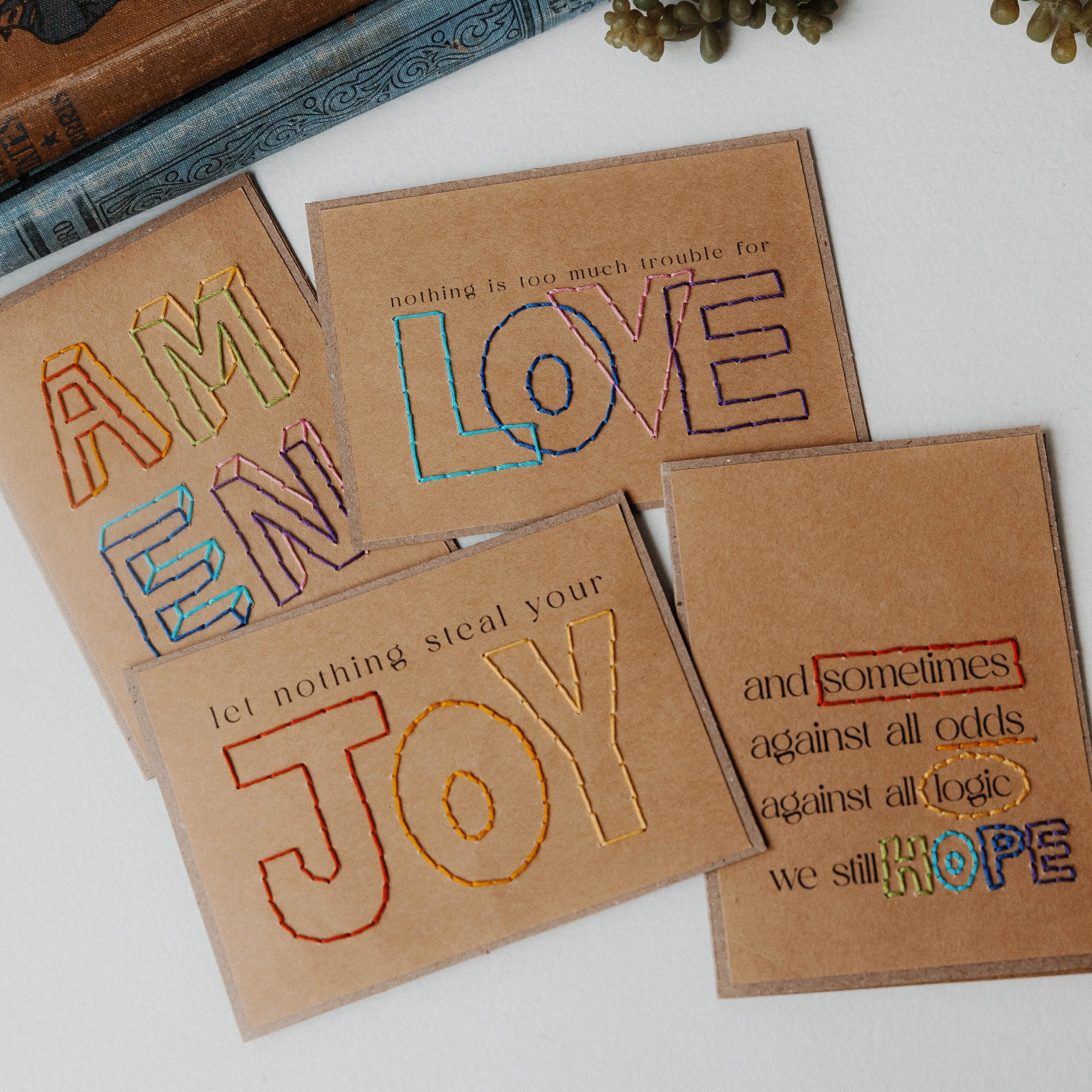 Four brown cards embroidered with colorful thread display positive messages, including AM EN, LOVE, let nothing steal your JOY, and a printed card about hope, surrounded by books and greenery.