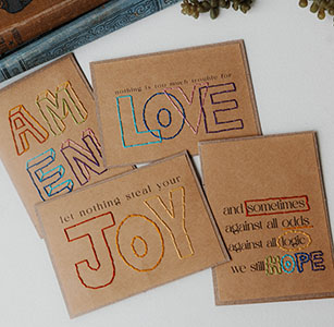 Four brown cards with motivational words stitched in colorful thread, including “AM EN,” “LOVE,” “JOY,” and “HOPE,” with additional positive phrases and decorative book and plant elements partly visible nearby.