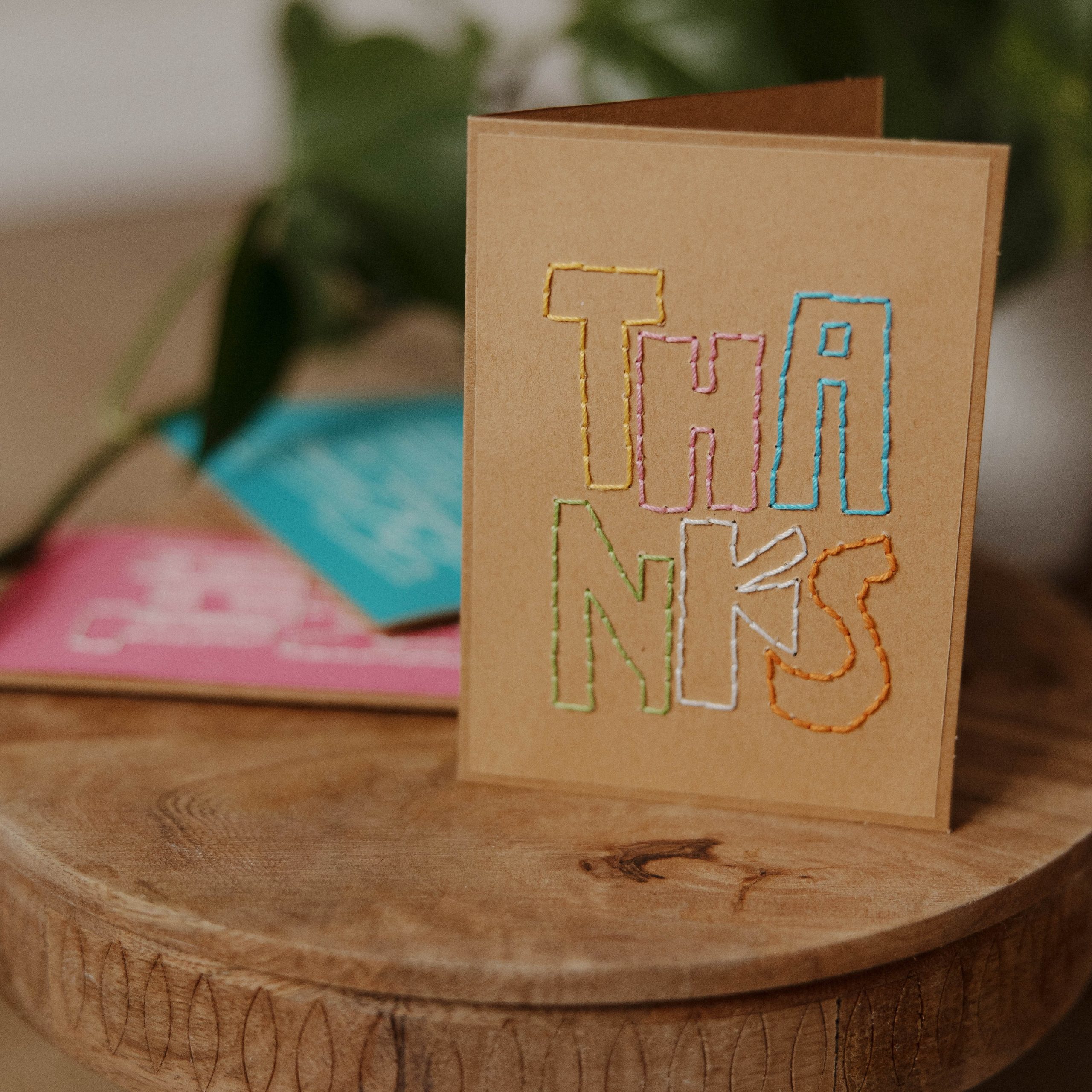 A brown card with the word THANKS stitched in colorful thread stands upright on a wooden surface. In the background, blurred green leaves and colorful cards are visible.