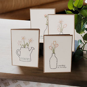 Four greeting cards with minimalist flower illustrations in vases and a watering can are displayed on a wooden surface next to a green plant. The cards feature simple line art with pastel-colored flowers.