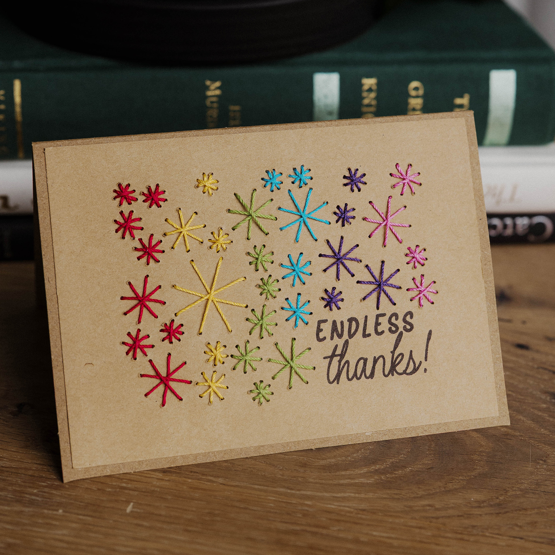A handmade thank you card decorated with colorful embroidered stars and the words “ENDLESS thanks!” written in black, propped up on a wooden surface in front of stacked books.